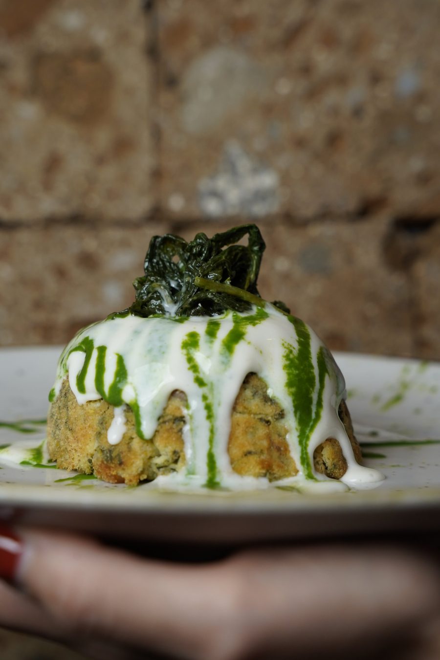 Sformato di verdure dorato ricoperto da salsa bianca e strisce di olio verde, visto di lato davanti a un muro di pietra