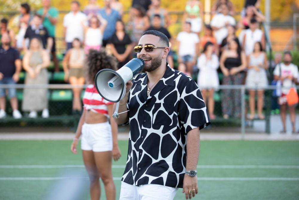 L'uomo in camicia maculata parla in un megafono sul campo, con una ballerina e la tribuna sfocate dietro