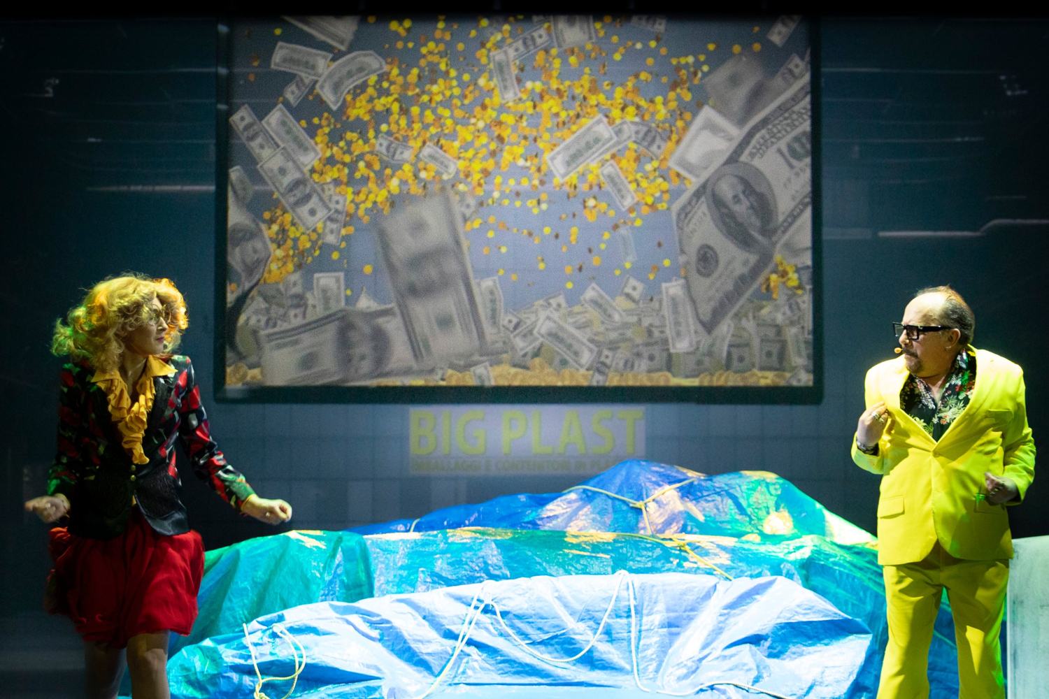 Una donna riccia e un uomo in completo giallo fluo ai lati del palco; sullo schermo piovono banconote e monete sopra un telo azzurro