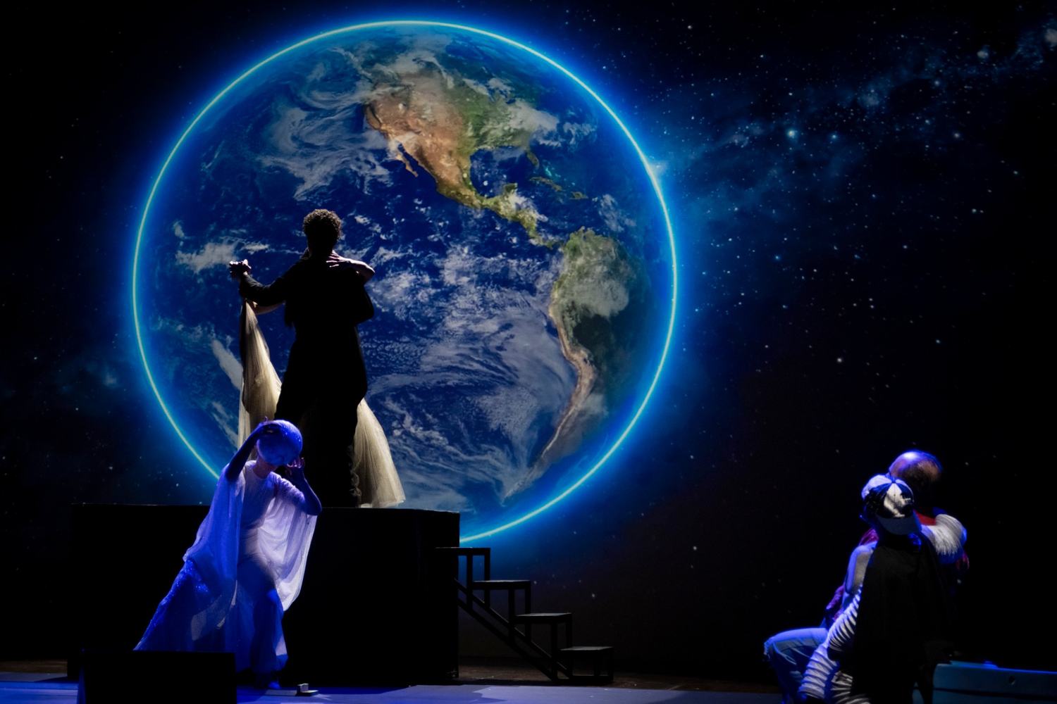 Sul palco, due figure ballano in silhouette davanti alla Terra proiettata; in basso a sinistra una figura in bianco regge la luna, a destra due persone si abbracciano