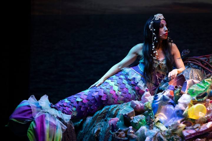 Una sirena dalla coda a scaglie viola e verdi, con perle tra i capelli, sdraiata su una montagna di plastica