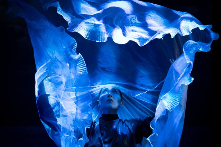 Una donna guarda verso l'alto sotto un grande velo azzurro luminoso che si increspa sopra di lei