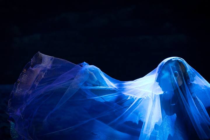 Una figura di profilo coperta da un velo trasparente blu, che si apre come un'onda nel buio
