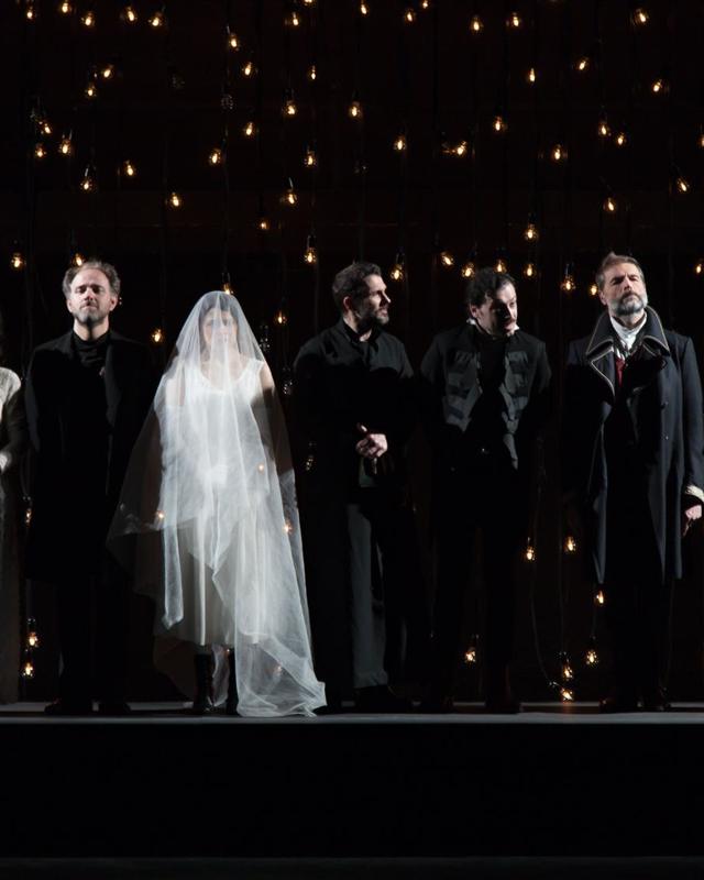Il cast in fila su un palco pieno di lampadine accese, con una sposa velata