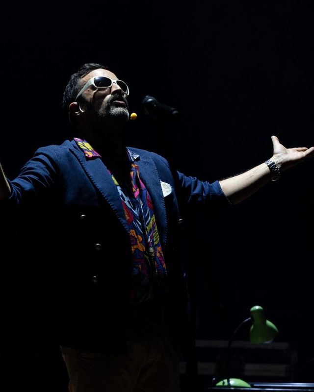 Un uomo con occhiali bianchi e giacca blu su camicia colorata allarga le braccia sul palco al buio