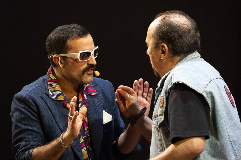Un uomo con occhiali bianchi da sole e giacca blu discute a mani aperte con un uomo con gilet di jeans, visto di spalle