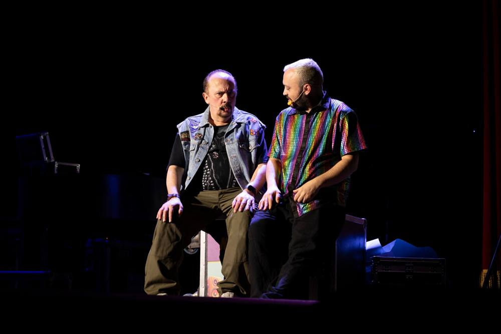 Due uomini seduti su una valigia da tour, uno con il gilet di jeans e l'altro con una camicia a paillettes arcobaleno, che gli parla