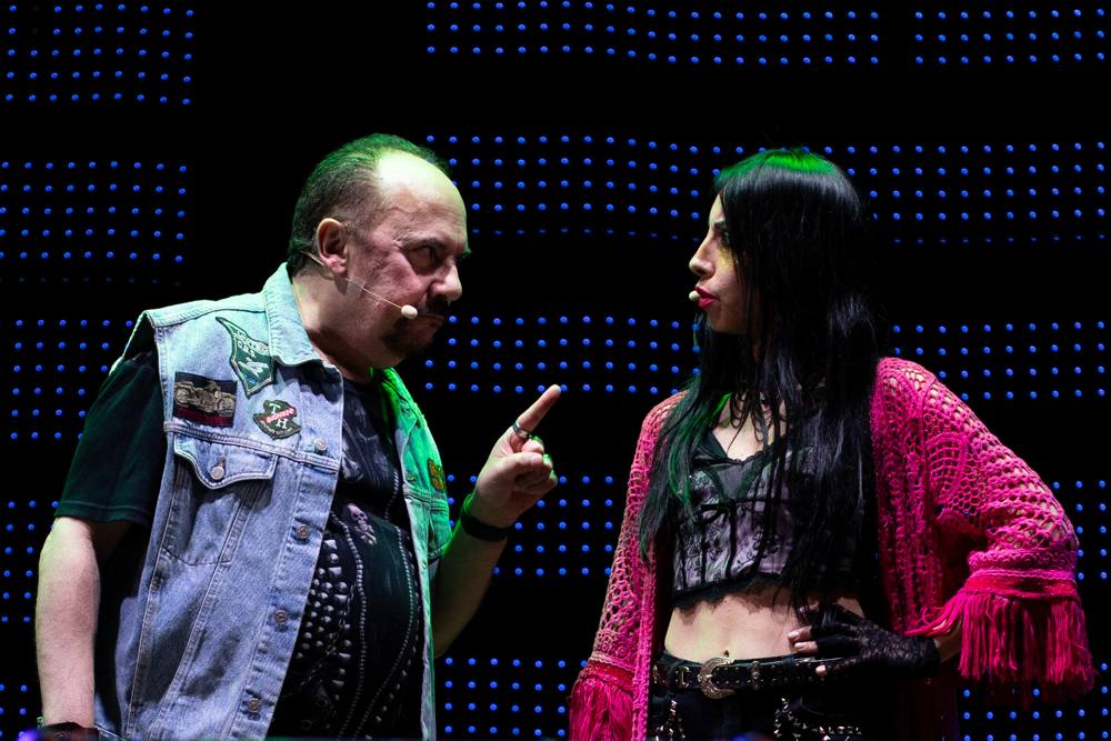 Un uomo con gilet di jeans punta il dito contro una donna con capelli neri e cardigan rosa, che lo guarda con una mano sul fianco