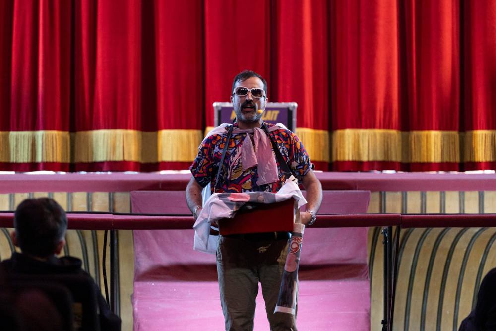 Un uomo con occhiali bianchi da sole, camicia colorata e una scatola rossa tra le mani, davanti al sipario rosso con frange dorate