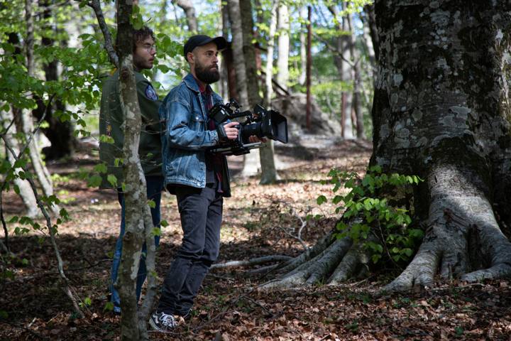 Un operatore con cappellino nero e giacca di jeans tiene la cinepresa nel bosco, accanto a un grande tronco; dietro di lui un ragazzo con gli occhiali