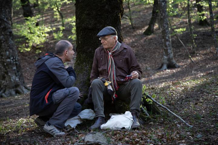 Due uomini, uno seduto contro un grande tronco e uno accovacciato, parlano a bassa voce